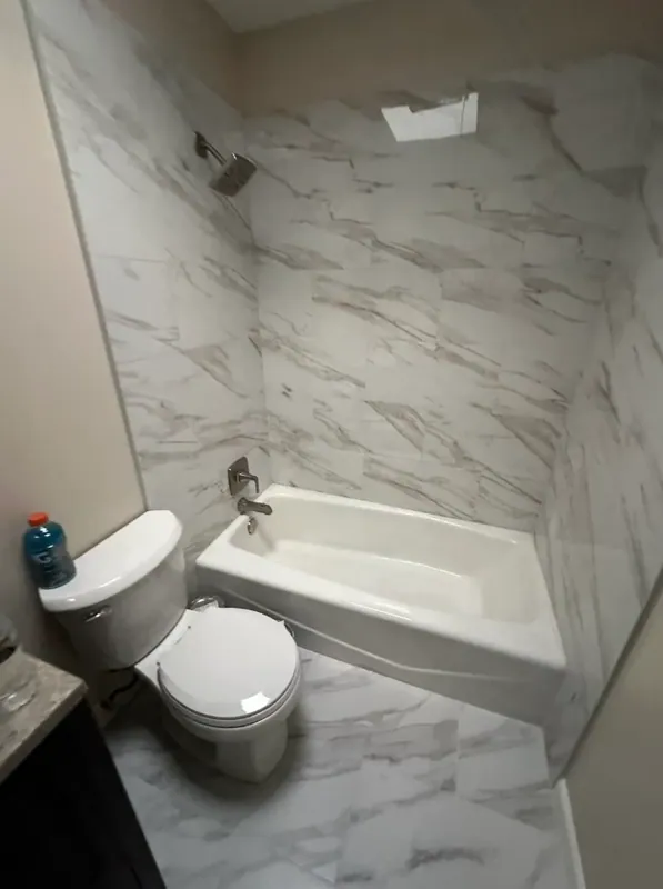 White tub alcove surrounded by gray-veined marble-look wall tile, with a white toilet and chrome shower fixtures.