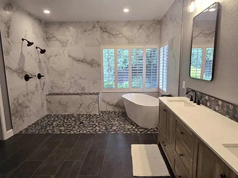 Bathroom with a wood-toned double vanity, dark rectangular floor tile, white soaking tub, rounded mosaic wet area, and marble-look wall tile.