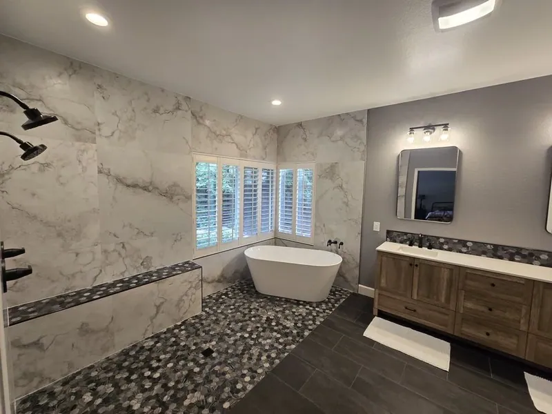 Second wide bathroom view showing black shower fittings, a rounded mosaic wet area, white soaking tub, dark floor tile, and wood-toned double vanity.