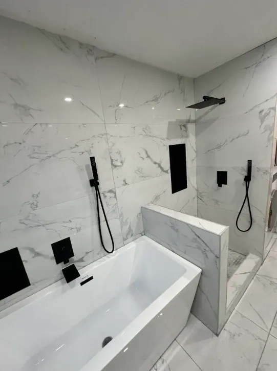 White soaking tub beside white marble-look wall tile, black shower fittings, and a low tiled partition.