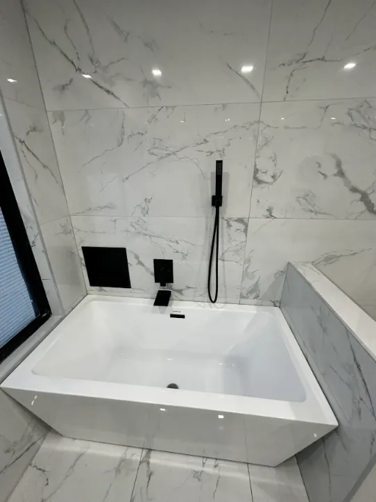 White soaking tub framed by gray-veined marble-look wall tile, a black hand shower, and a black-trimmed window.