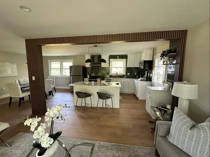 Medium-brown plank-style floor running through a living area toward a white kitchen framed by dark wood slats.