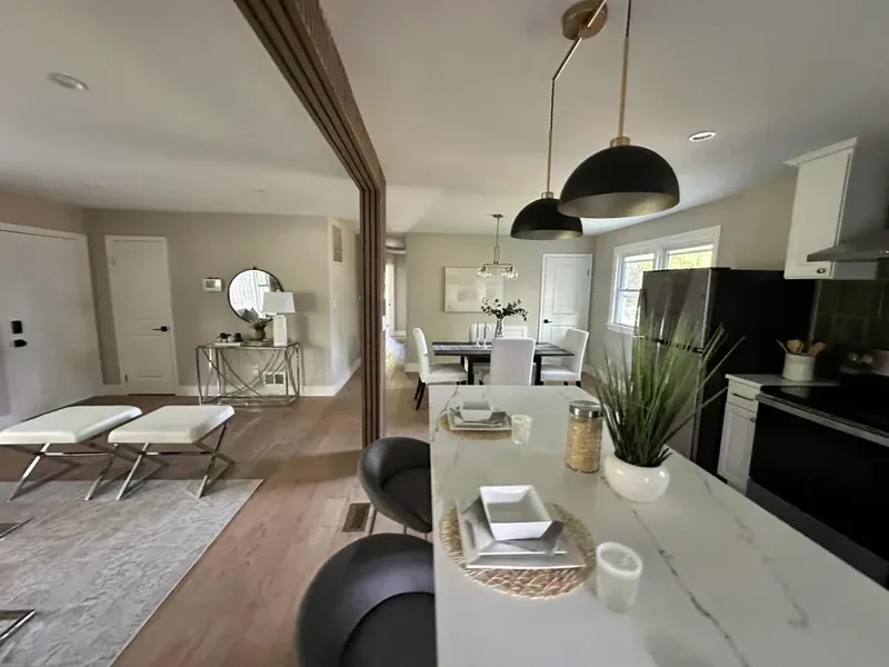 White kitchen peninsula with black pendant lights, black backsplash, and medium-brown plank-style flooring extending toward dining and living areas.